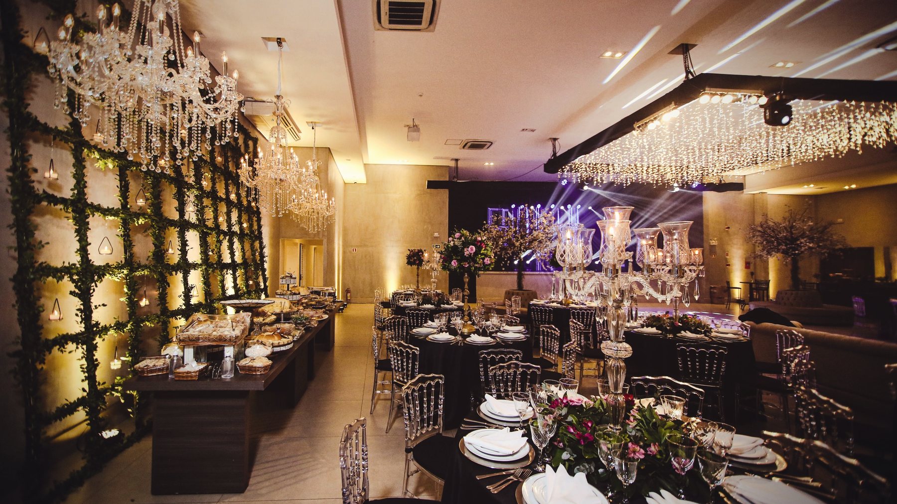 Salón de eventos montado de noche, con arañas de cristal, manteles oscuros y luz dorada.